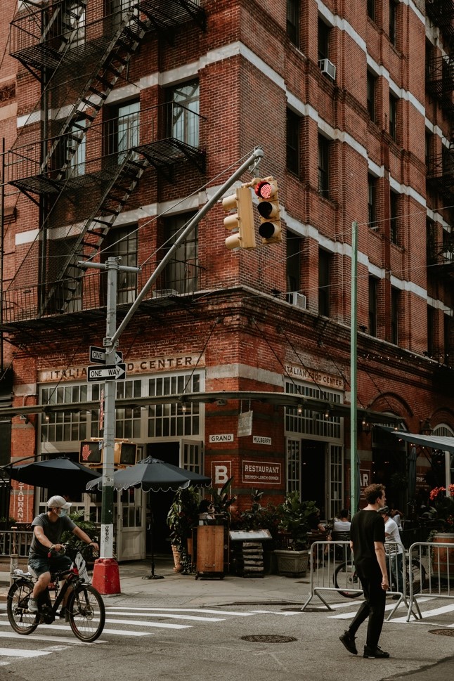 Immeuble new yorkais dans un quartier style industriel à New York, il y a une route avec un cycliste, des passants traversant des passages piétons. Il y a un feu de circulation jaune au centre de l'image et des escaliers en métal sur les bâtiments.